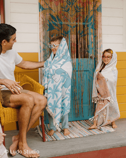Two children wrapped in patterned towels standing on a porch with a person sitting, Ludo Studio watermark visible.