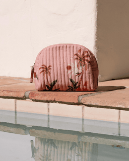 Pink quilted pouch with palm tree design on a wooden ledge by a pool.