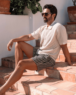 Man sitting on stone steps wearing a patterned shirt and shorts, with plants in the background.