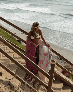 Woman standing on a staircase by the beach with a pink flamingo towel draped over her