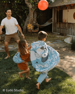 Two children and a man playing outdoors with patterned cloaks, Ludo Studio watermark visible.