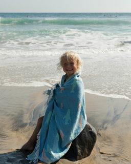Little girl wrapped in the blue swirling shark print towel, sitting on a rock at the beach.