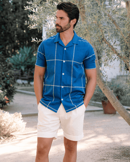 Man wearing a blue checkered shirt and white shorts standing outdoors.
