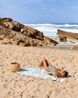 Woman lying on a towel on a sandy beach with ocean and rocks in the background