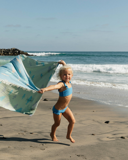 Little girl on the beach holding up a light green swirling shark print towel, waves in the background.