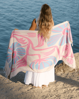 Woman sitting on a beach with a pink and blue patterned towel draped over her shoulders.