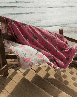 Pink party blanket with black flamingo pattern draped over a wooden rail by the beach.