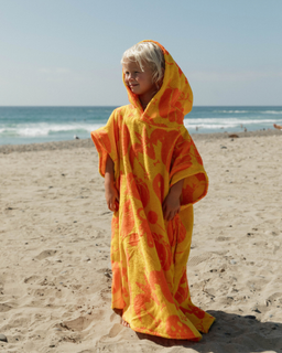 A young child wearing a bright yellow and orange patterned hooded beach towel or poncho stands on a sandy beach with the ocean and blue sky in the background.