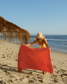 Woman on a beach holding a red towel with wavy pattern, under a straw umbrella.