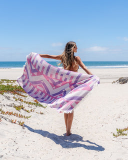 A woman stands on a sandy beach holding a large patterned beach towel or sarong spread wide in front of her. The textile has a geometric chevron pattern in pink, purple, and white with fringe on the edges.