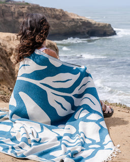 A woman sits on a sandy beach facing the ocean, wrapped in a large blue and white patterned blanket or throw. The blanket features an abstract leaf-like design in teal blue and white, with fringed edges visible on the right side.