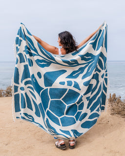 A woman standing on a sandy beach holds up a large woven blanket with a blue sea turtle pattern on a white background.