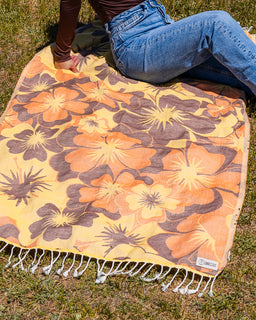 A person sitting on a yellow and brown floral blanket with white fringe, laid out on green grass outdoors.