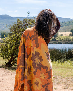 A person standing outdoors in front of a lake and hills, wrapped in a lightweight orange and yellow floral blanket or wrap with large brown flower motifs.