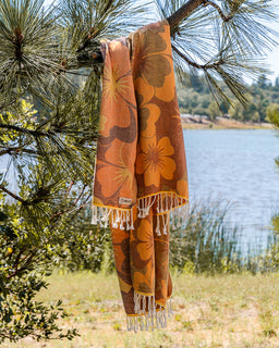 A patterned orange and brown towel with white tassels hanging from a pine tree branch, with a lake and grassy shoreline in the background.