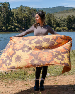 A woman standing outdoors by a lake holds up a large patterned towel with oversized floral motifs in orange, brown, and cream. Trees, water, and hills are visible in the background.