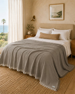 A styled bedroom scene showing a bed made with a textured taupe-gray crinkle blanket, white sheets, and a tan accent pillow. The room has warm beige walls, a window with sheer curtains and a view of the ocean, and a woven jute rug.