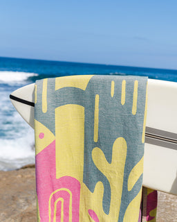 A colorful beach towel with a yellow, pink, and slate blue abstract pattern draped over the side of a white surfboard, with the ocean and blue sky in the background.