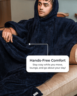 A man wearing a plush navy blue hooded blanket poncho sits on a beige leather couch. A white text box overlay reads 'Hands-Free Comfort Stay cozy while you move, lounge, and go about your day!'.
