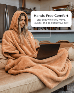 A woman sits on a beige sofa wrapped in a plush tan hooded blanket, using a laptop. A white text bubble reads, 'Hands-Free Comfort Stay cozy while you move, lounge, and go about your day!' The setting is a modern living room with a kitchen in the background.