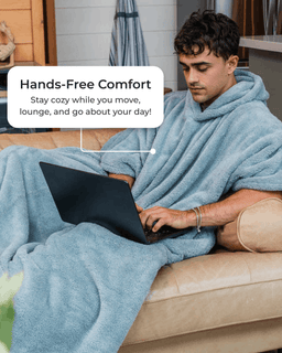 A man lounging on a beige couch wrapped in a light blue plush fleece blanket, using a black laptop. A white text bubble reads 'Hands-Free Comfort Stay cozy while you move, lounge, and go about your day!'.