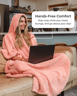 A woman sits on a beige couch wearing a soft peach-pink hooded fleece blanket robe while using a laptop. A white text bubble reads 'Hands-Free Comfort Stay cozy while you move, lounge, and go about your day!' The setting is a real indoor living room environment.