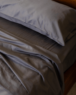 Bed with grey sheets and a white pillow on a wooden bed frame.