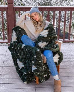 Girl on with snowy trees behind her wearing a beanie  and sitting on a chair and wrapped up in a green christmas tree print drift blanket