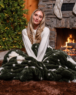 Woman sitting in front of a fireplace and christmas tree in a cozy cabin wrapped up in Christmas tree drift blanket
