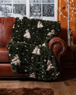 Christmas tree print blanket draped over a brown sofa with snowy trees in the background through the window