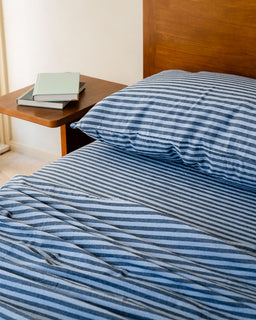 Bed with blue striped bedding and a wooden headboard, side table with books in the background.