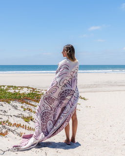 A woman standing on a sandy beach wrapped in a large patterned towel with purple and white swirl motifs, with the ocean and blue sky behind her.