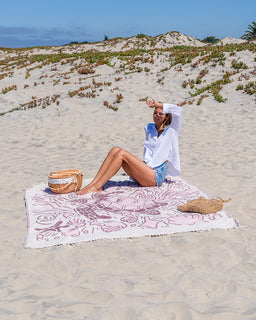A woman relaxes on a large white beach towel with pink floral patterns on a sandy beach. She is wearing a white button-up shirt and denim shorts, with a woven straw bag beside her on the sand.