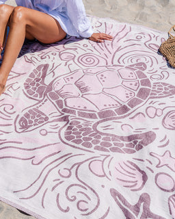 A large white beach towel with a pink turtle and wave pattern, spread out on sand with a person sitting on one corner of it.