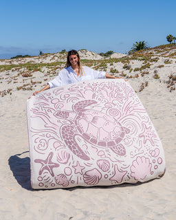 A woman stands on a sandy beach holding up a large square beach towel or blanket featuring a pink sea turtle and ocean-themed line art on a white background.