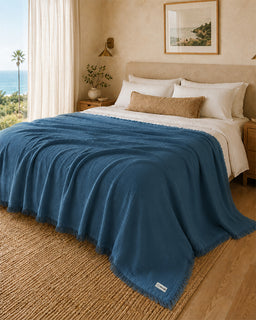A textured blue throw blanket with fringe is draped over a bed in a warm, sunlit bedroom with a beach view, beige walls, and wood flooring.