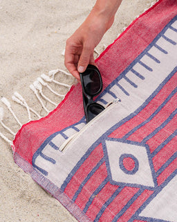 Person holding sunglasses over a red and blue patterned towel on sand