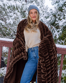 Woman wearing a beanie wrapped in a chocolate colored blanket with snowy trees in the background