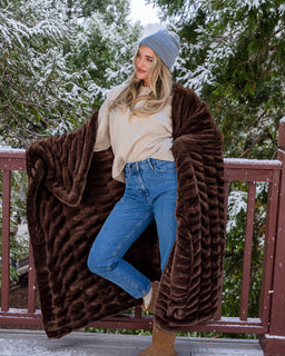 A woman stands on a snowy porch draped in a large, plush, ribbed brown throw blanket. She is wearing a gray beanie, a cream sweater, blue jeans, and brown boots.