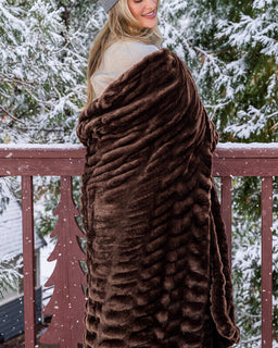 A woman wrapped in a plush, ribbed brown faux fur blanket standing on a wooden deck railing with snow-covered pine trees in the background.
