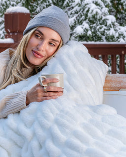 Girl on a cabin deck with snowy trees in the background with cozy snow colored drift blanket holding a mug