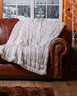 White cozy drfit blanket draped over couch with a snowy trees in the background through a window