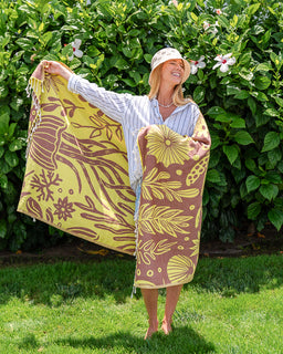 A woman standing outdoors in a garden holds up a large tropical-patterned towel or wrap. The fabric is primarily yellow and brown with botanical motifs like leaves, flowers, and seashells.