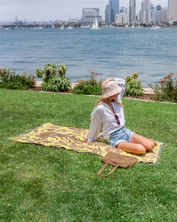 A woman sits on a yellow and brown patterned blanket on a green lawn, with a body of water and city skyline in the background. A woven straw bag rests beside her.