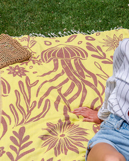 A yellow outdoor blanket with a brown jellyfish and floral pattern, laid on green grass. A person wearing a white pinstriped shirt and denim shorts is sitting on the blanket, with a woven straw bag partially visible on the left.