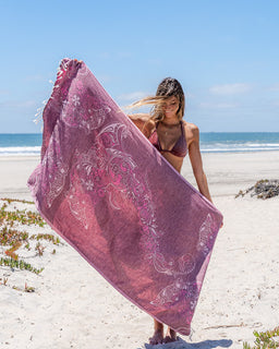A woman on a sandy beach holds up a large, semi-sheer pink sarong with white paisley patterns, with the ocean and blue sky in the background.