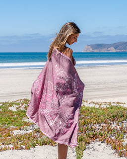 A woman standing on a sandy beach wrapped in a dusty pink sarong with white floral and paisley patterns, with the ocean and blue sky in the background.