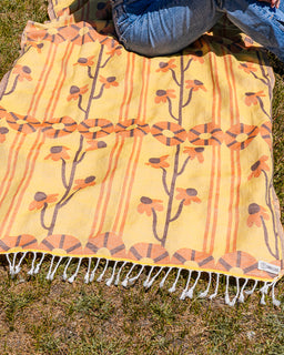 A yellow woven blanket with orange and brown floral patterns and white fringe, laid out on grass with a person's blue jeans visible at the top edge.