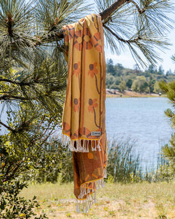A mustard yellow woven scarf with orange and brown floral patterns and white fringe, hanging from a pine tree branch with a lake and forest in the background.