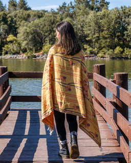 A woman walking on a wooden dock by a lake, viewed from behind, wearing a yellow patterned towel with orange and brown accents.
